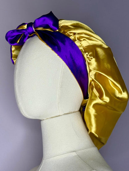 AQueenzDen gold and purple double layer satin bonnet with adjustable ties and a bow detail.