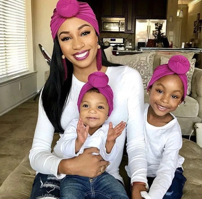 AQueenzDen Mommy and Me matching set: a woman and two children wear matching magenta cotton top knot turbans and white long-sleeved shirts.